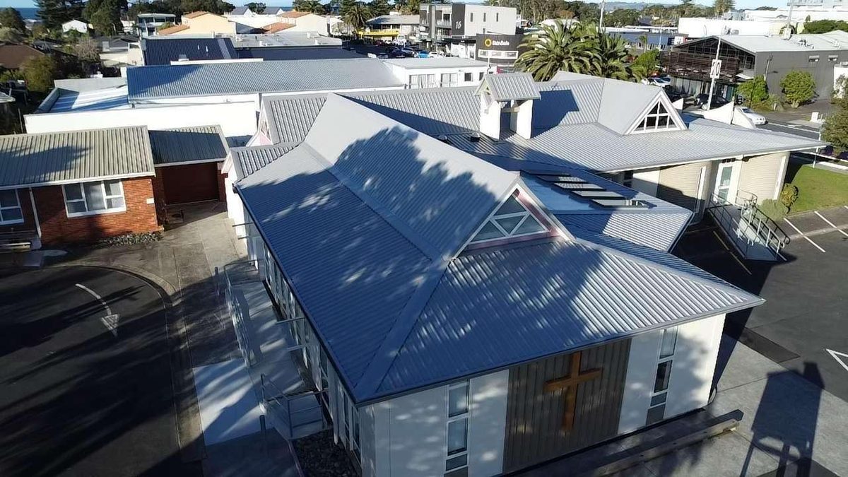 Drone view of a church with a large grey longrun steel roof, a gable window and a cross on the end wall