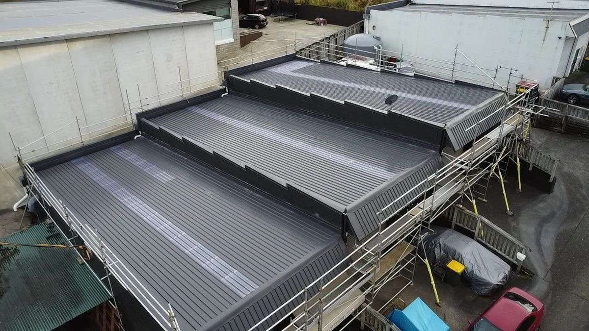 Dark grey longrun roof in three stepped sections on a commercial building, with scaffolding and edge protection