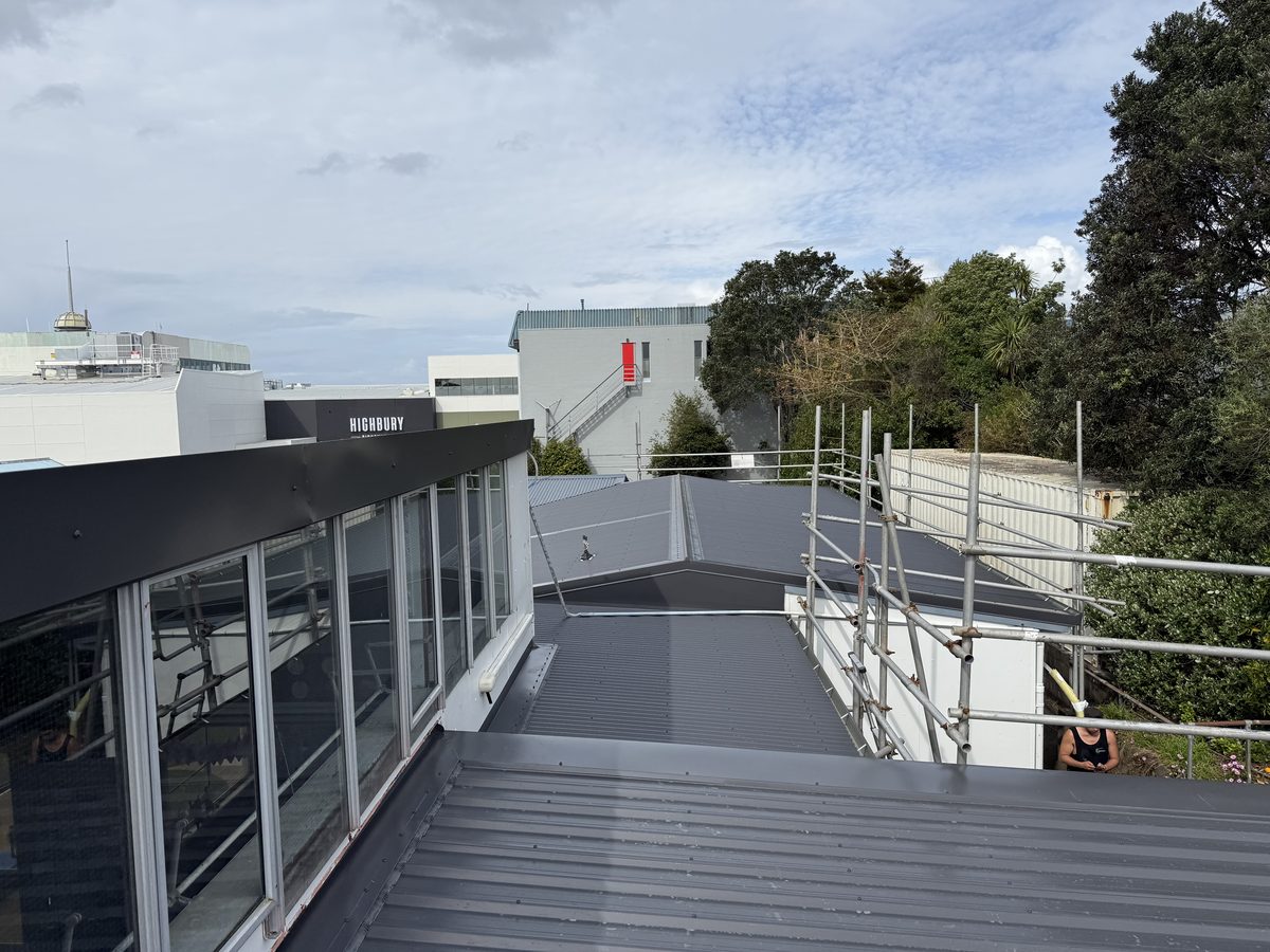 Dark grey longrun steel roof beside a glazed upper storey, with scaffolding along one edge