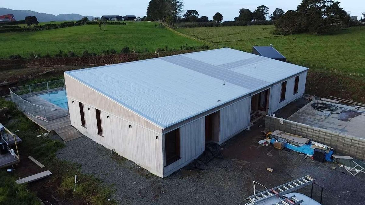 Aerial view of a rural home with a light grey low pitch roof beside a fenced pool, farmland behind