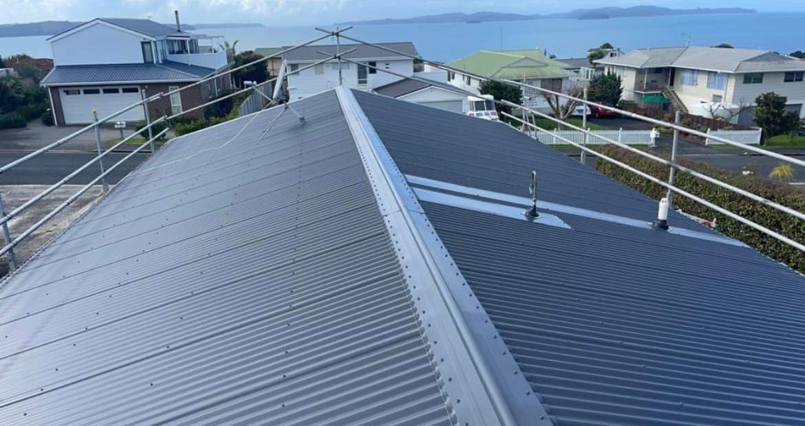 View along the ridge of a dark blue-grey corrugated steel roof towards houses and the sea