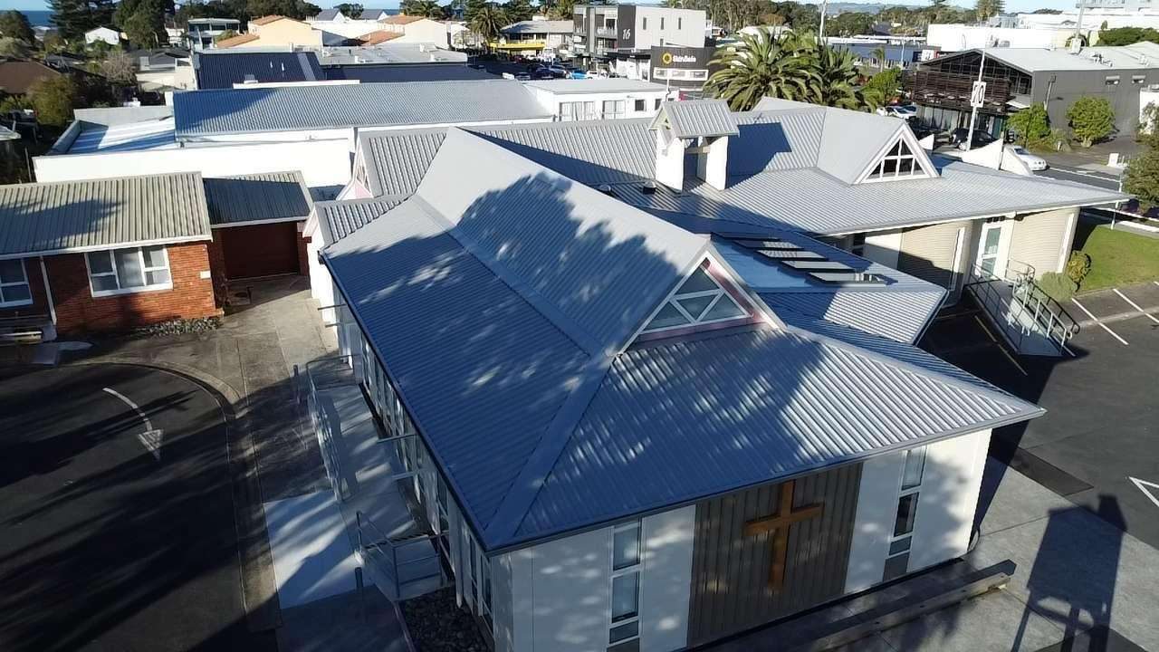 Drone view of a church with a large grey longrun steel roof, a gable window and a cross on the end wall