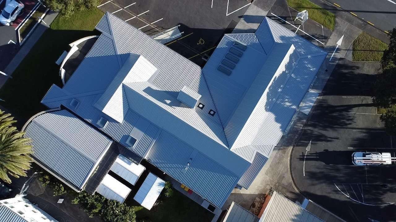 Drone photograph looking straight down on a large light grey corrugated roof with hips, valleys and skylights, beside a car park