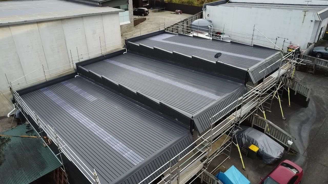 Dark grey longrun roof in three stepped sections on a commercial building, with scaffolding and edge protection