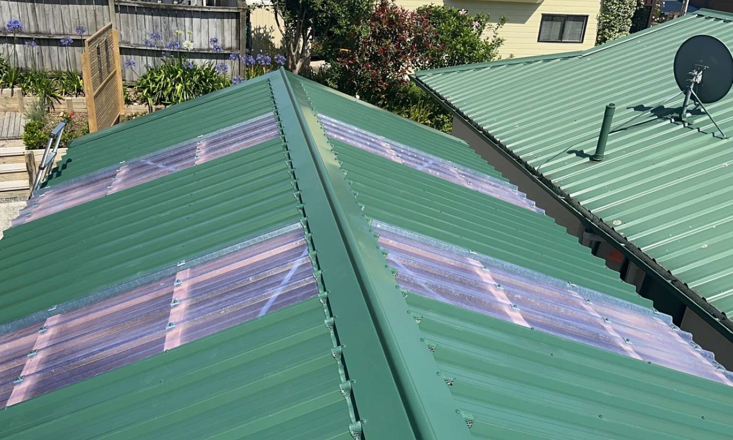 Dark green corrugated steel roof with clear roofing panels either side of the ridge, a satellite dish and gardens behind