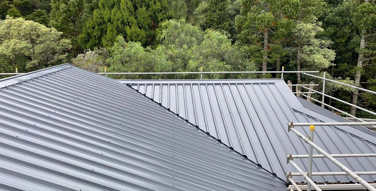 Two planes of a mid grey ribbed steel roof meeting at a valley, with scaffolding and bush behind