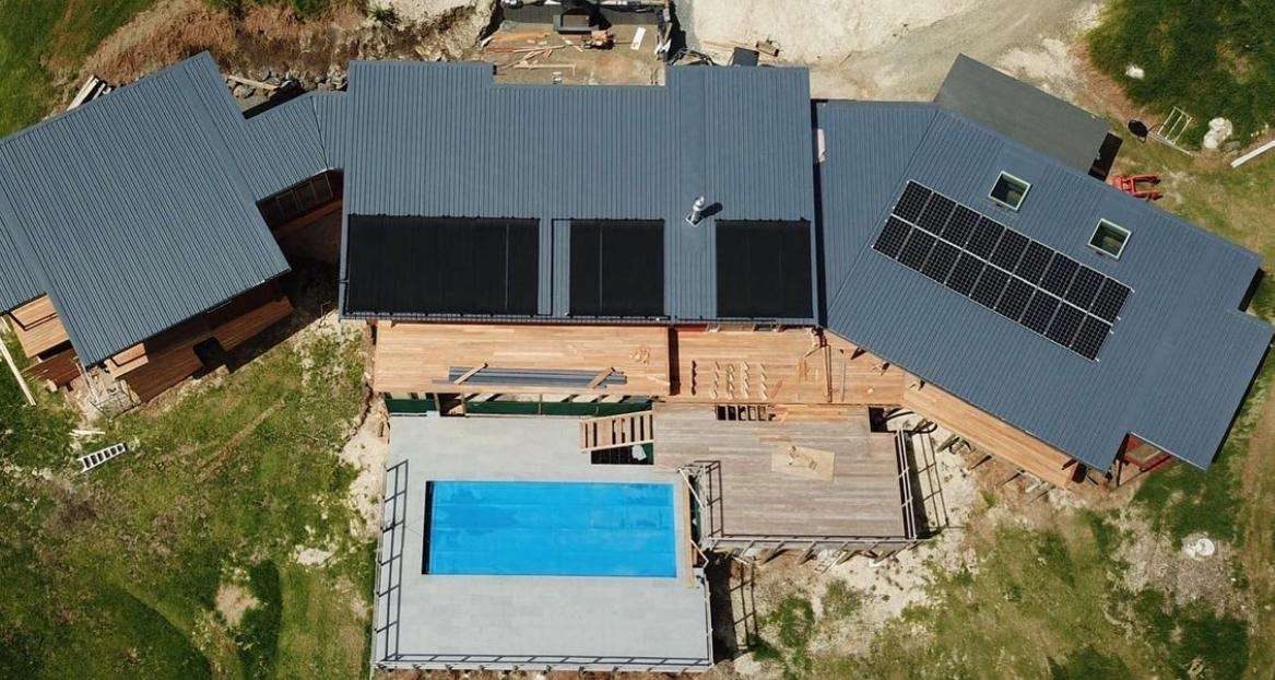 Aerial view of a new timber-clad home with dark grey longrun roofing, solar panels and a swimming pool