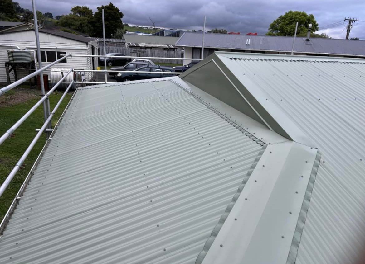 Pale sage green corrugated steel roof with hip capping running up to a small gablet, a scaffold rail along the edge