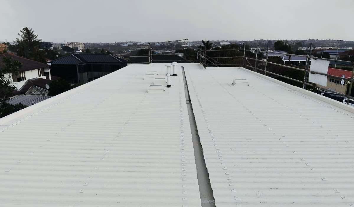Wide white longrun steel roof with edge protection on a commercial building, looking out over a suburb
