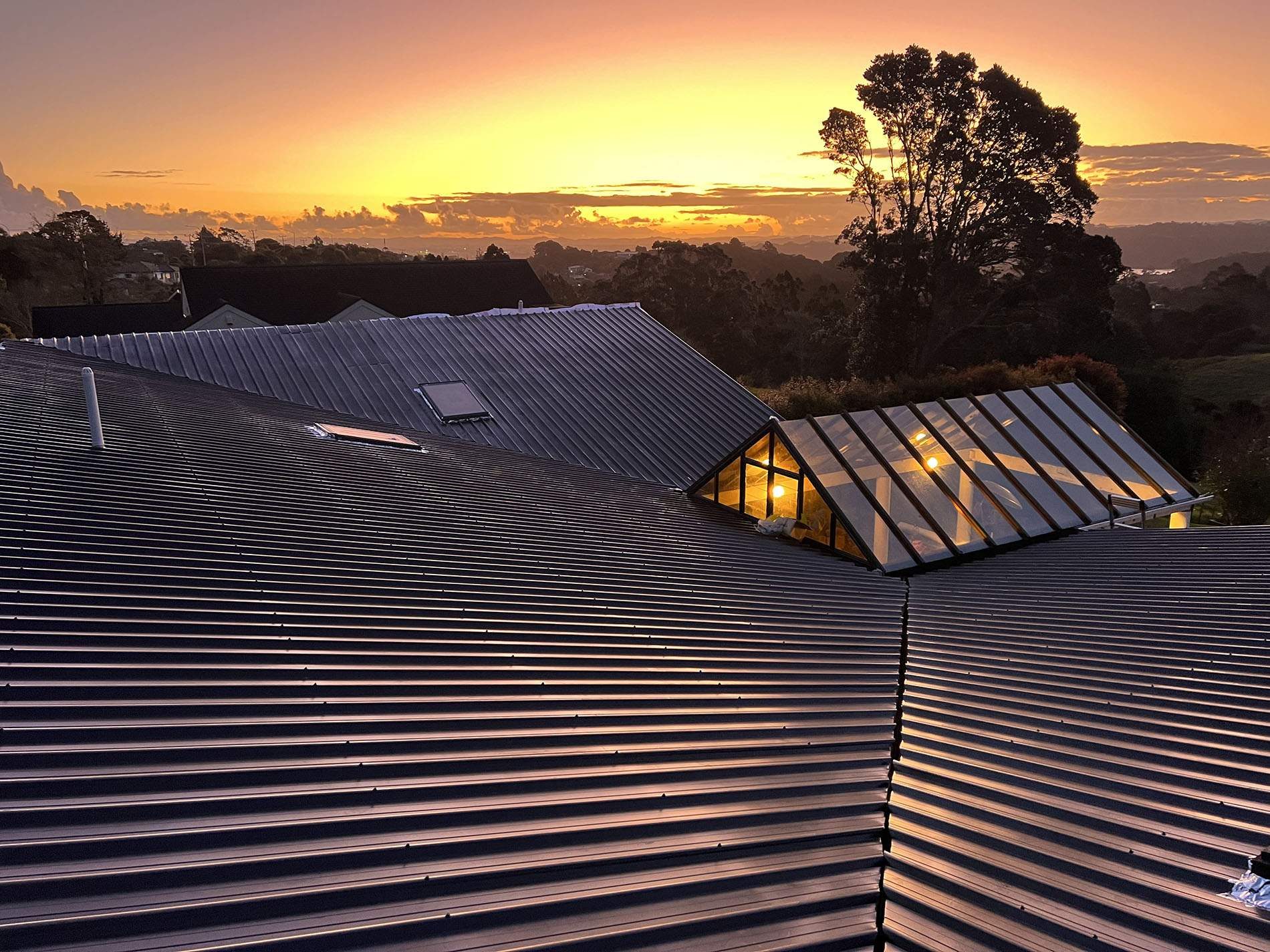 Dark ribbed steel roof with a valley and skylights at sunset, a glazed conservatory roof lit from inside