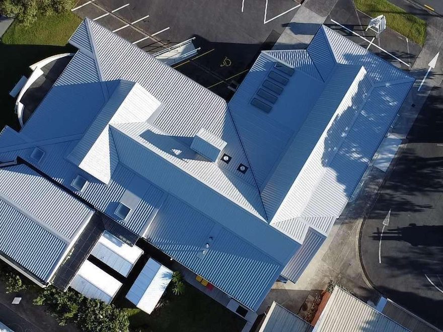Drone photograph looking straight down on a large light grey corrugated roof with hips, valleys and skylights, beside a car park
